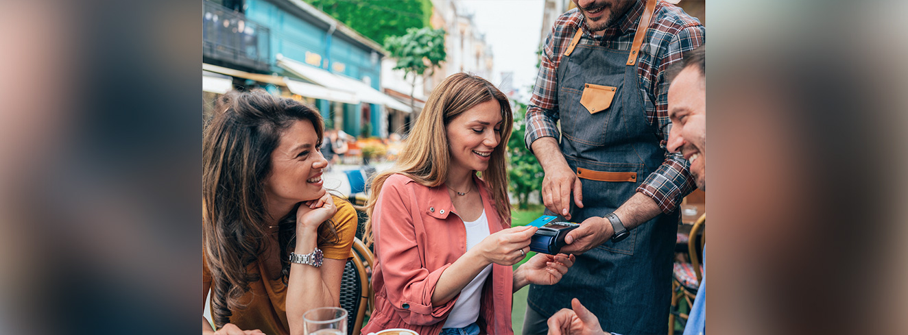 Gäste bezahlen mit Karte im Außenbereich eines Cafés während ein Service-Mitarbeiter ein Kartenlesegerät hält